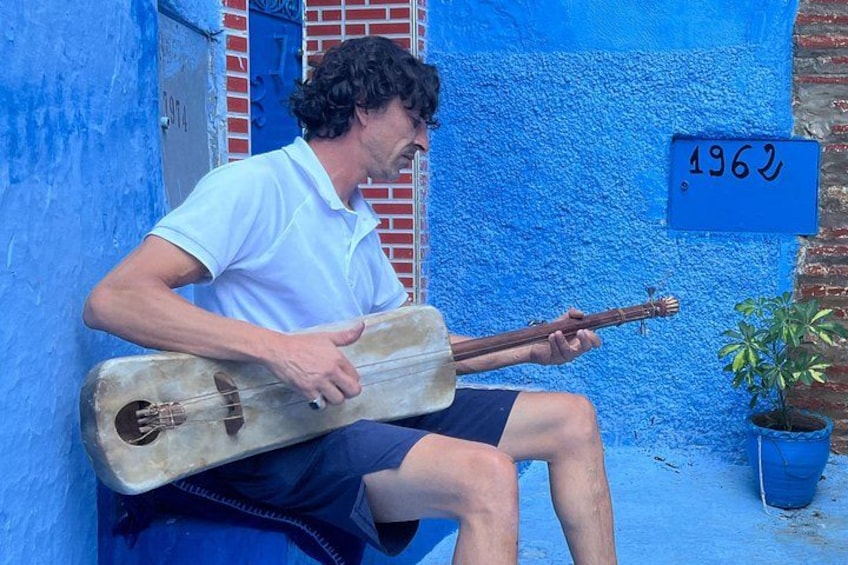 A person playing a traditional stringed instrument, sitting on a blue wall with a blue door and a number sign. 1