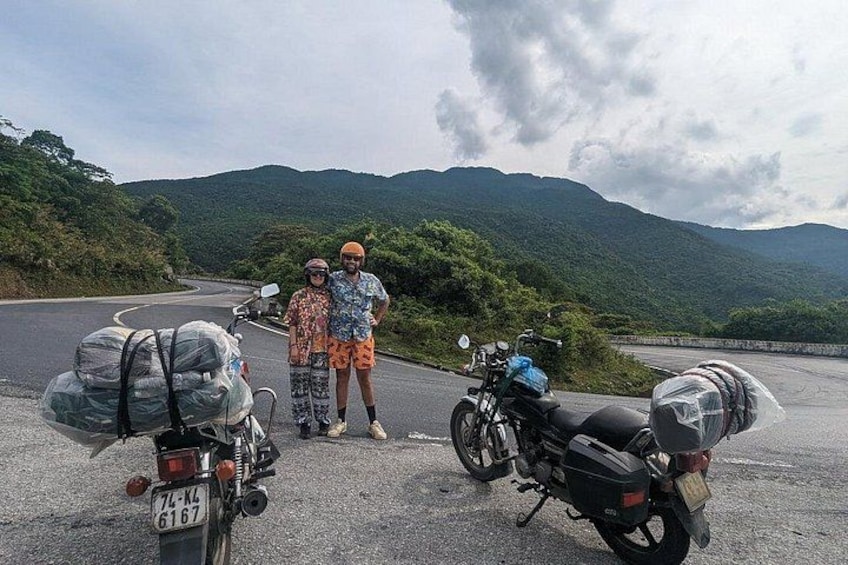 Zwei Motorräder stehen an einer Straße, im Hintergrund eine gebirgige Landschaft. 2