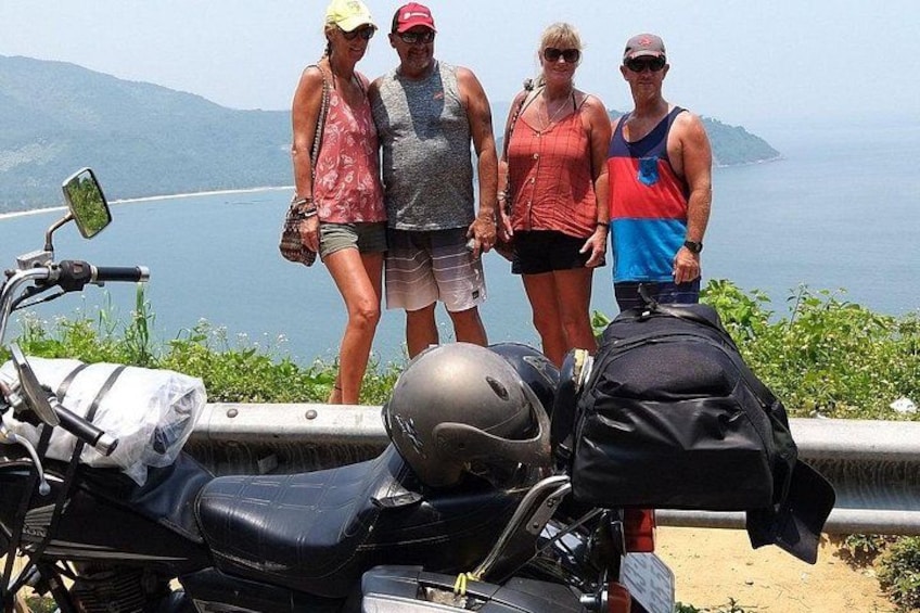 Eine Gruppe von Menschen steht an einer Küstenstraße neben einem Motorrad, im Hintergrund Meer und Berge. 4