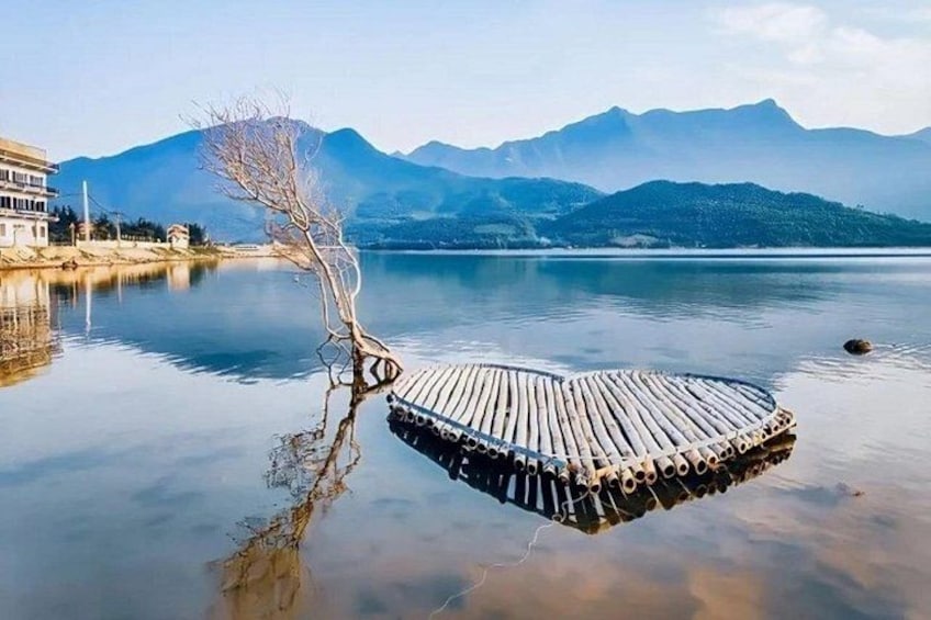 Ein Floß aus Holz auf ruhigem Wasser, ein kahlstehender Baum und Berge im Hintergrund. 5
