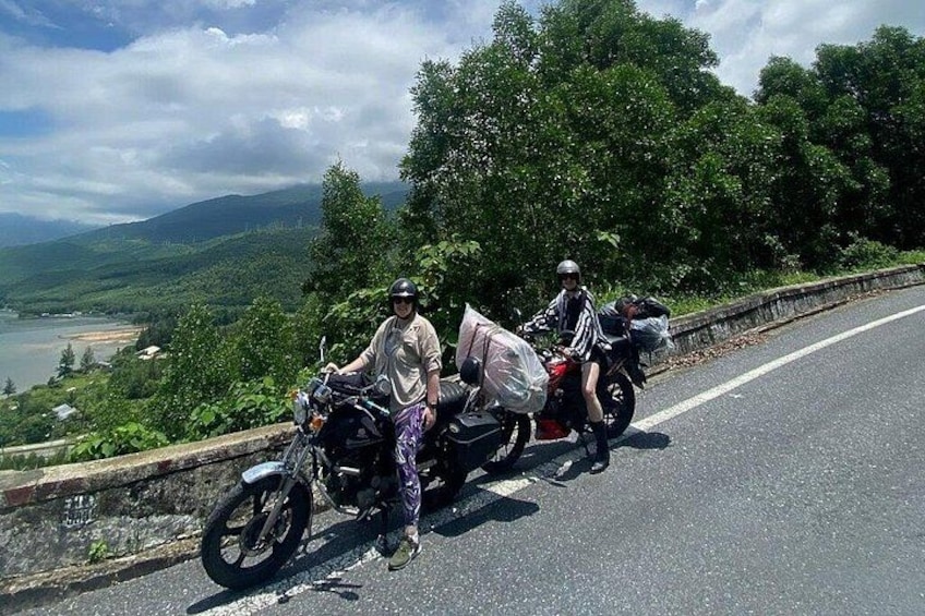 Zwei Motorradfahrer auf einer Straße mit herrlicher Aussicht auf einen See und Hügel im Hintergrund. 1