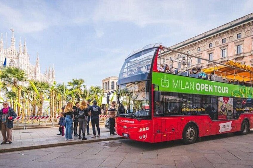 A red double-decker bus with 'Milan Open Tour' written on it, parked on a city street with palm trees and historic buildings in the background. 4