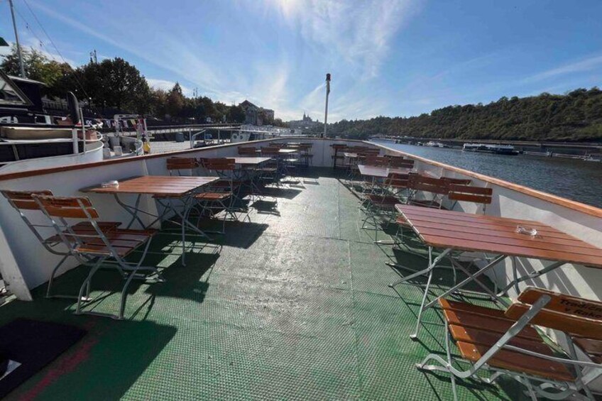 A boat deck with tables and chairs, a green floor, and a river with a bridge in the background. 2