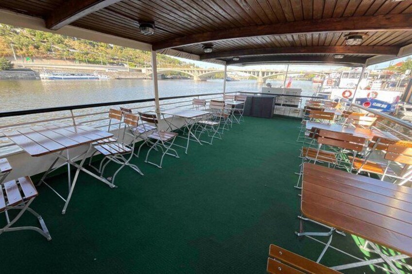 A covered outdoor seating area with tables and chairs, overlooking a river with boats and a bridge. 3