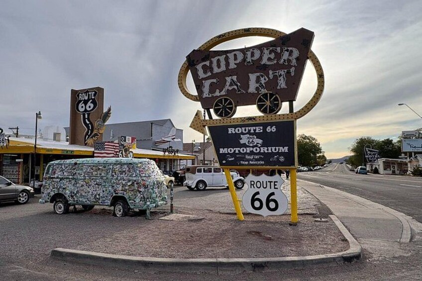 Ein Schild der historischen Route 66 mit einem 'Copper Cart'-Schild, einem Verdeckwagen und einer Motorradrennbahn. 2