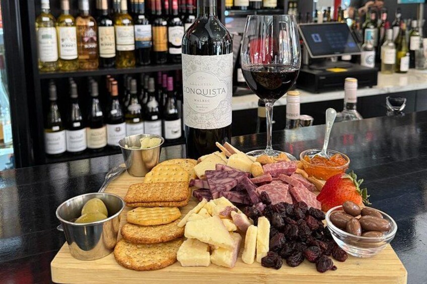 En skærebræt med forskellige oste, pålæg, frugter og kiks, ledsaget af en flaske Conquist vin og et glas rødvin. 2
