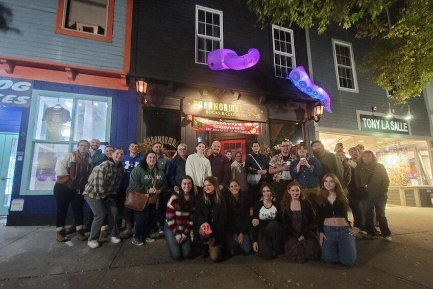 A group of people posing in front of a building with a neon sign and a storefront. 1