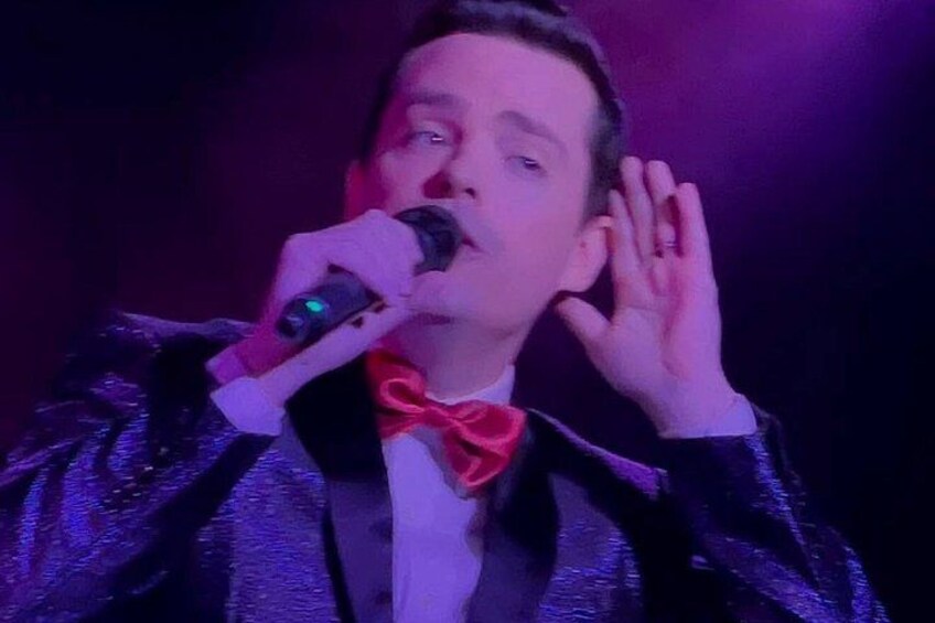 Une personne portant une veste noire brillante et un nœud papillon rouge, et tenant un microphone. 4