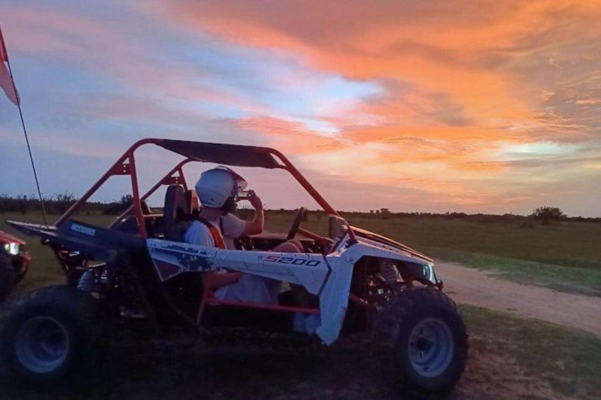 A 4x4 vehicle with a driver and passenger, parked on a dirt path during sunset. 1