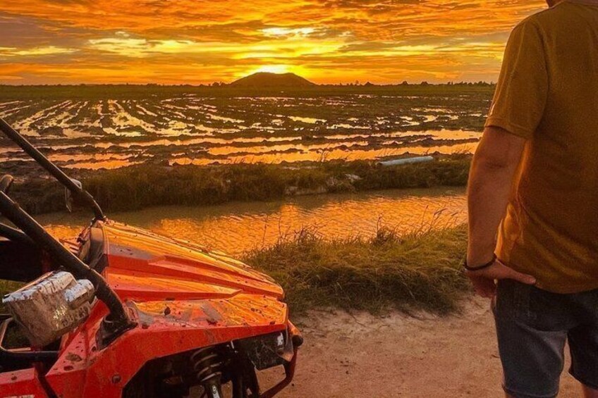 A person standing by a red ATV on a dirt path with a sunset over a flooded field. 3