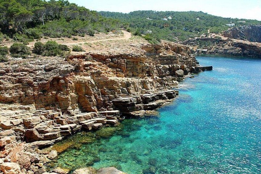 A coastal cliff with clear blue water, rocky terrain, and sparse vegetation. 3