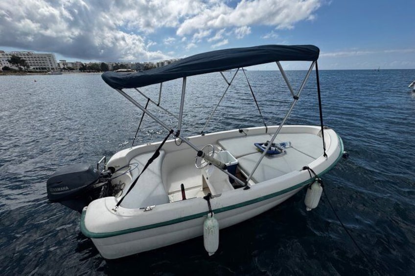 A small motorboat with a canopy, docked at a calm body of water with a clear sky and distant buildings. 4