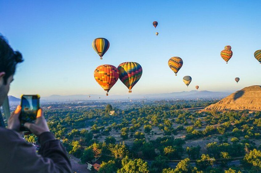 Iemand maakt een foto van luchtballonnen boven een landschap met bomen en een heuvel. 1