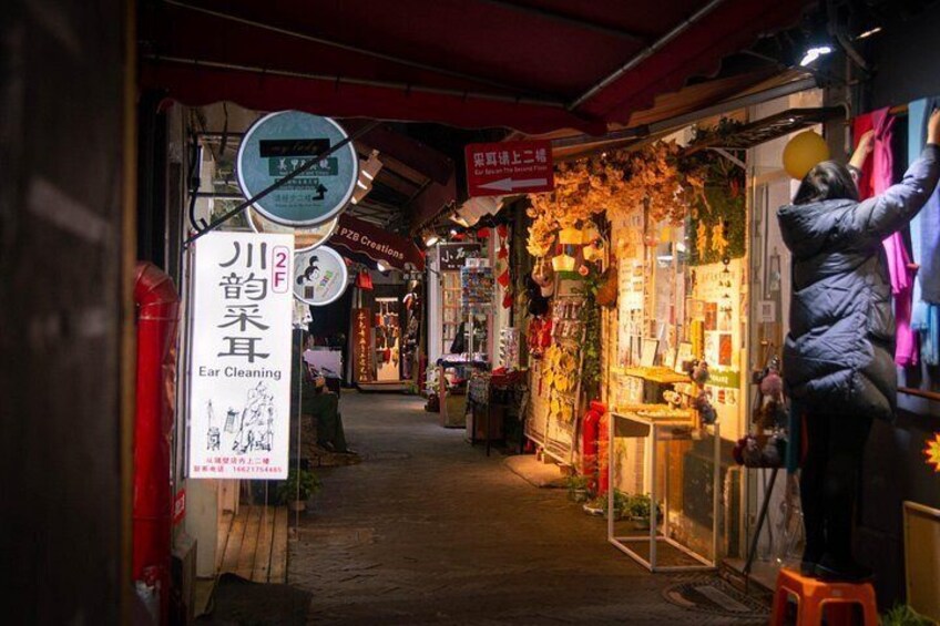 A narrow alley with shops on either side, a sign for ear cleaning, and a person reaching for something on a shelf. 1