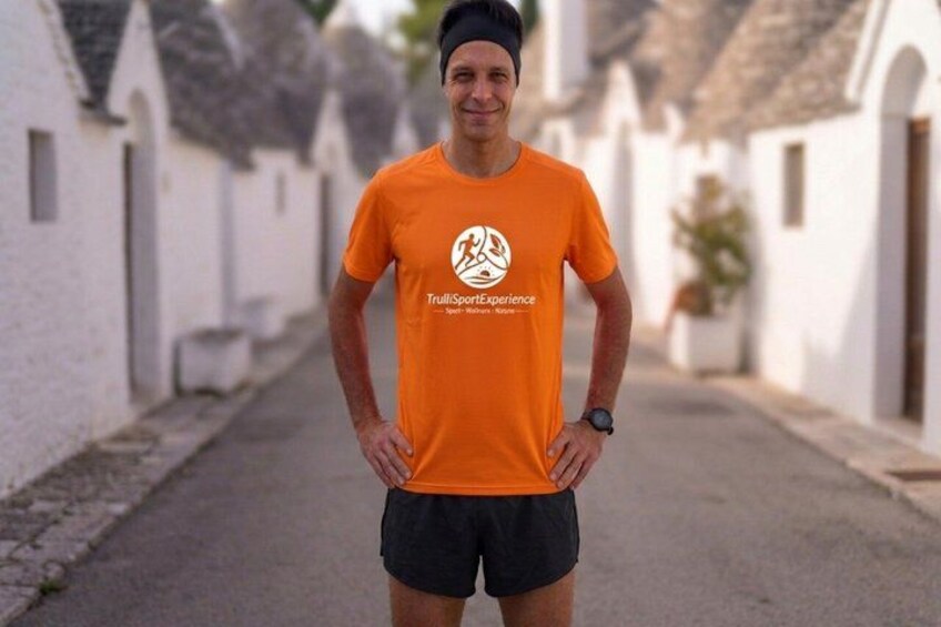 Een persoon met een oranje T-shirt met een logo, staand op een straat met witte gebouwen en een geplaveid pad. 2