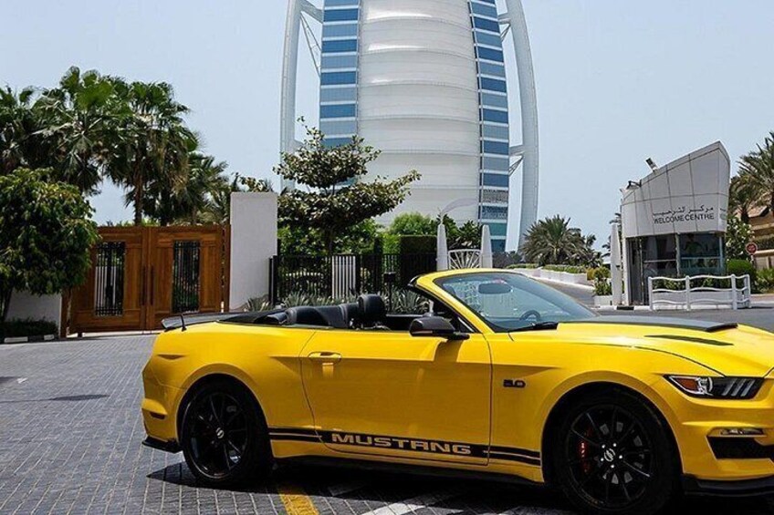 A yellow Mustang convertible parked in front of a modern building with a sail-like structure. 2