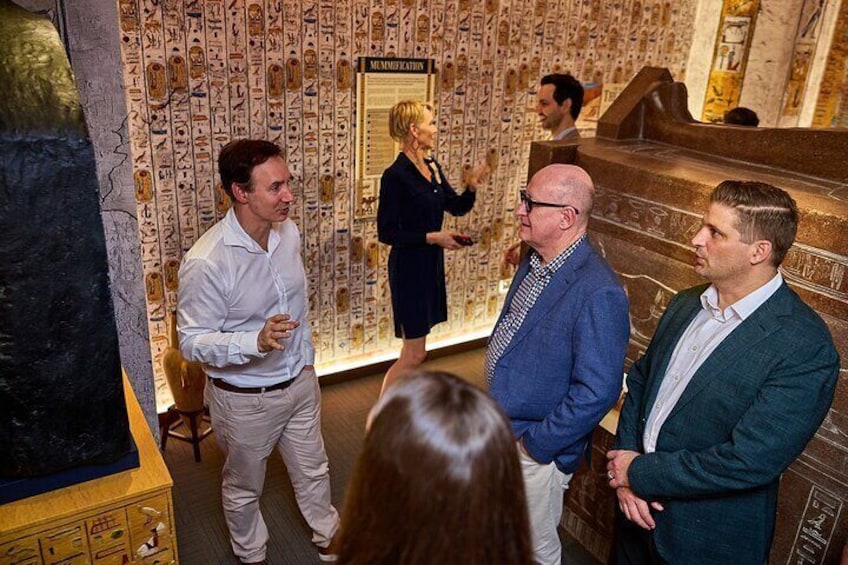 A group of people in an indoor setting with hieroglyphic wall decorations and a stone structure resembling a sarcophagus. 1