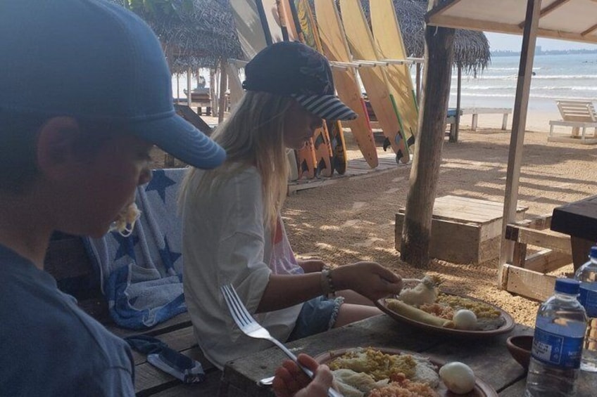 Personas cenando en una mesa al aire libre con vistas a la playa, incluyendo una estructura de techo de paja y tablas de surf. 3