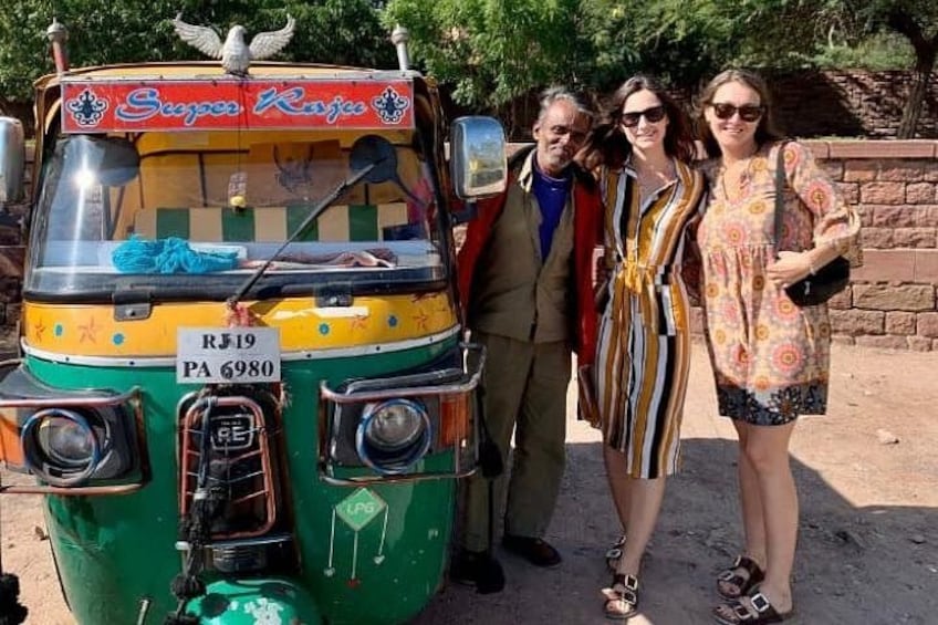 A colorful auto-rickshaw with a license plate, parked on a dirt ground with people standing beside it. 2