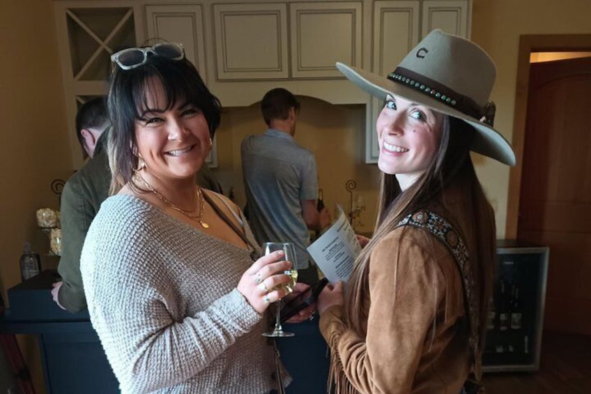 To personer med drikkevarer, den ene iført en lys sweater og den anden en cowboyhat, i et rum med et skab og en dør i baggrunden. 4