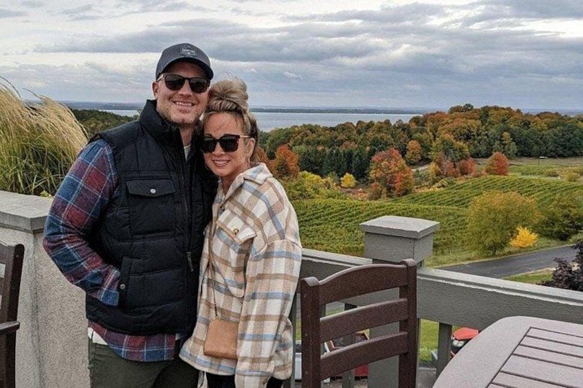 A couple standing on a balcony overlooking a scenic landscape with trees and a body of water. 4
