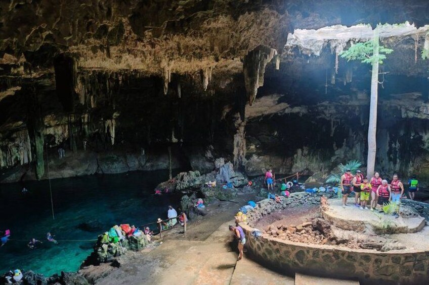A cave with stalactites, a water pool, and people exploring. 3