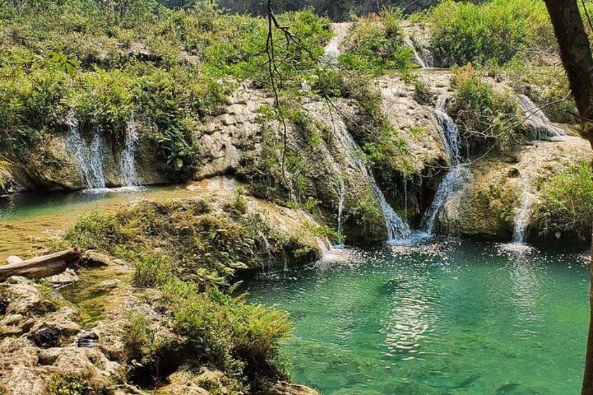 Ein Wasserfall stürzt über Felsen in einen klaren Pool, umgeben von viel Grün. 4