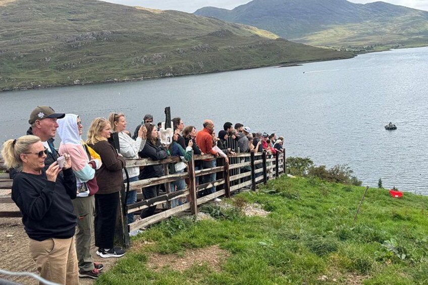 Un groupe de personnes se tient près d’une clôture en bois, d’où l’on aperçoit un lac et, au loin, des collines. 4