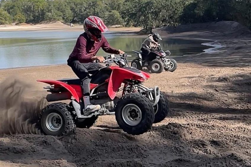 A person riding a red quad bike on a sandy beach with a lake in the background. 1