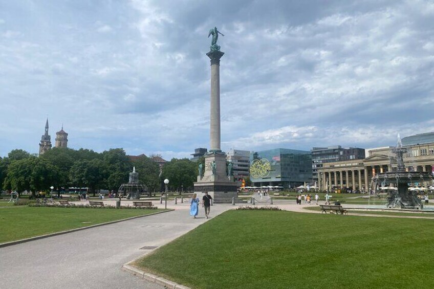 A park with a tall monument, a fountain, and a statue. There are buildings in the background and a tower. 5