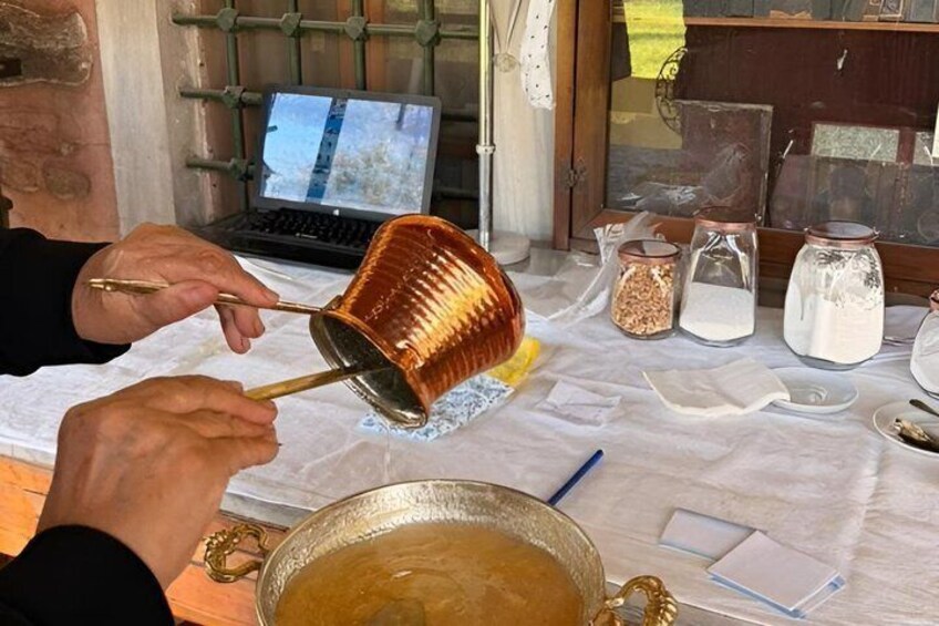 A person pouring liquid from a copper pot into a glass container on a table with various jars and a laptop. 1