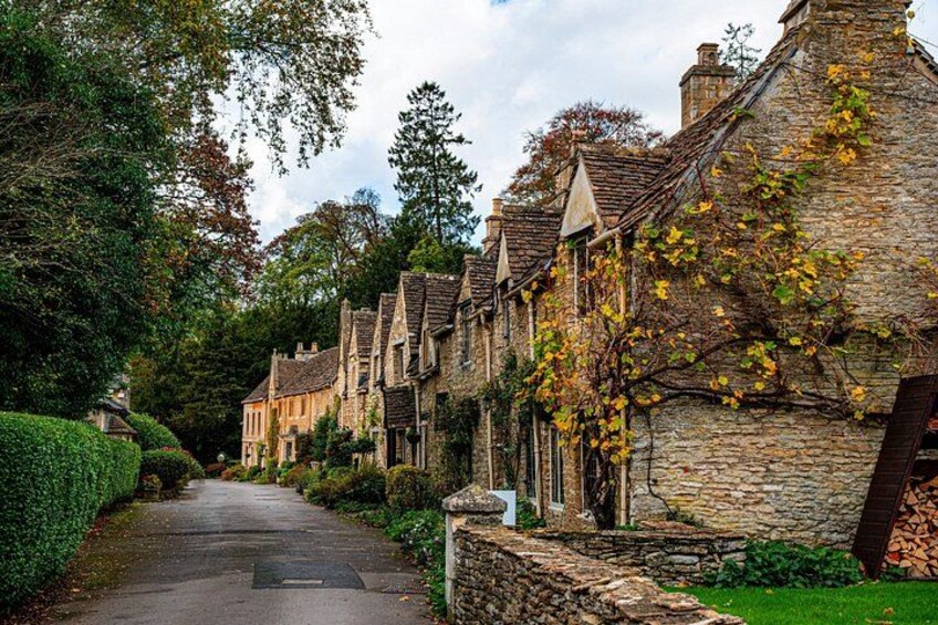 A narrow street lined with stone houses, a hedge on one side, and a tree with yellow leaves on the other. 2