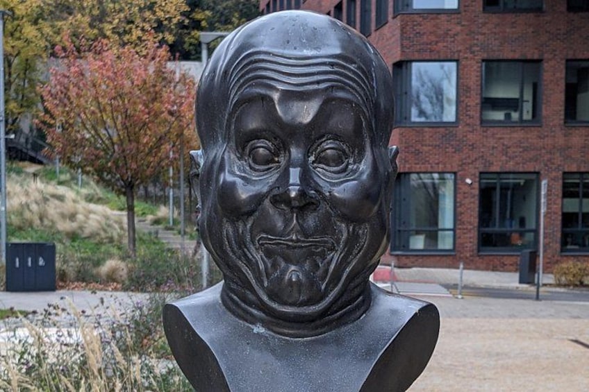 Una scultura raffigurante un volto in uno spazio pubblico, con un edificio e alberi sullo sfondo. 1