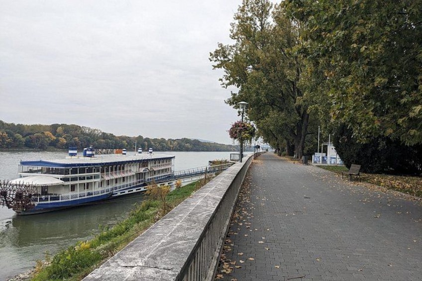 Un fiume con una barca ormeggiata a sinistra, un viale lastricato lungo la sponda e alberi che costeggiano il percorso. 5
