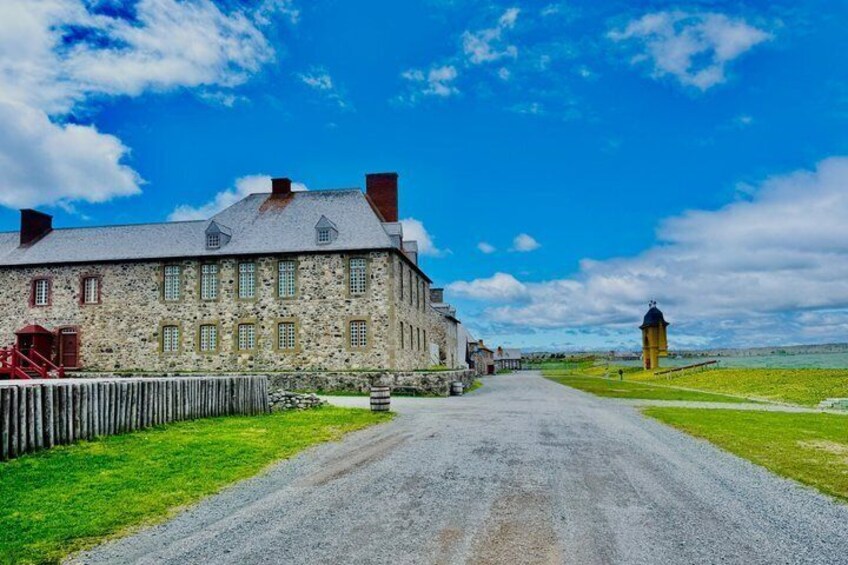 A stone building with a gravel road leading to it, a wooden fence, and a tower in the distance. 3
