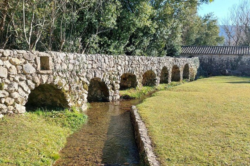 Un puente de piedra con arcos sobre un estrecho canal, rodeado de hierba y árboles. 2