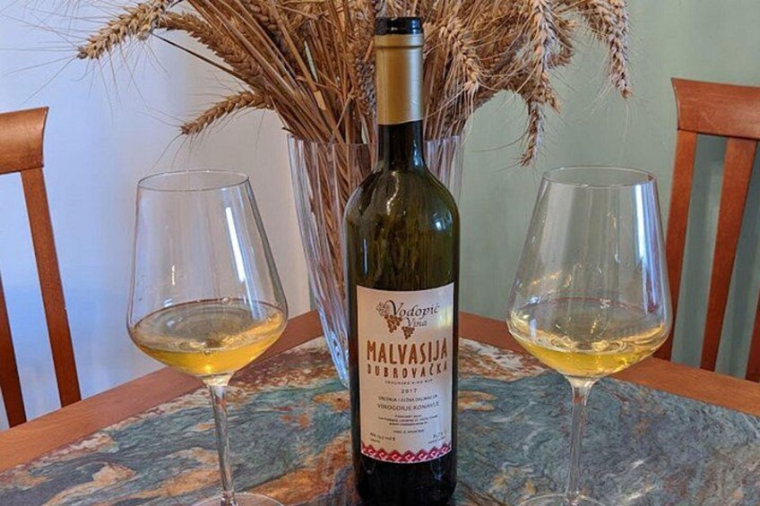 Una botella de vino Malvasija, dos copas de vino y un jarrón con espigas de trigo sobre una mesa. 3