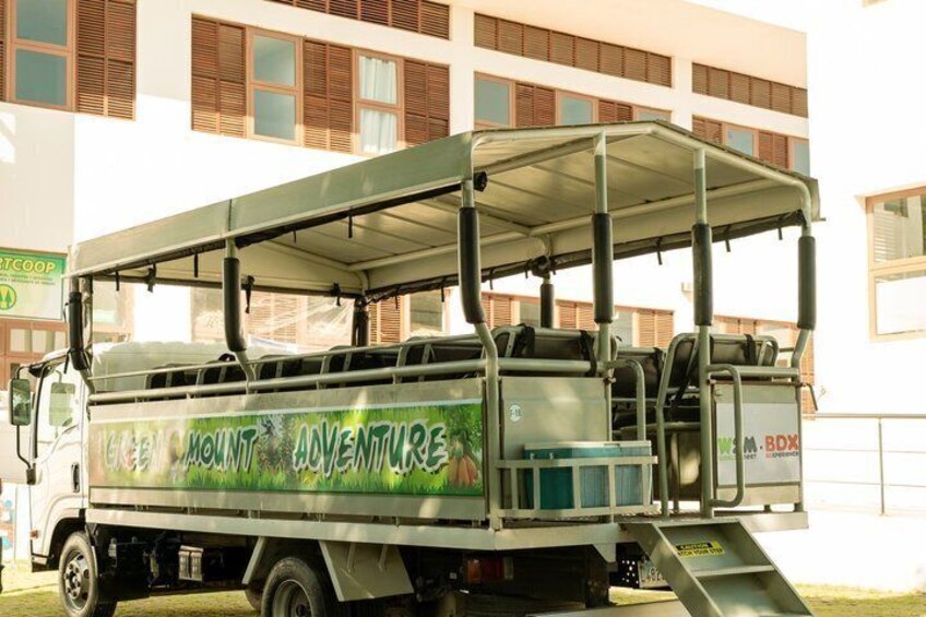 Een groen en wit safari voertuig met 'Mount Adventure' op de zijkant, geparkeerd voor een gebouw met ramen en een bord waarop 'M. BOX' staat. 2