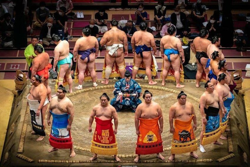 Un grupo de luchadores de sumo de pie en círculo, algunos con mawashi tradicionales y otros con mawashi azules y anaranjados. 1