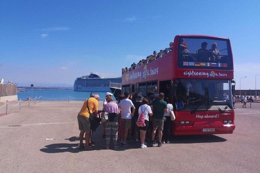 Un autobús de dos pisos rojo con pasajeros subiendo, un crucero al fondo y un cielo azul despejado. 4