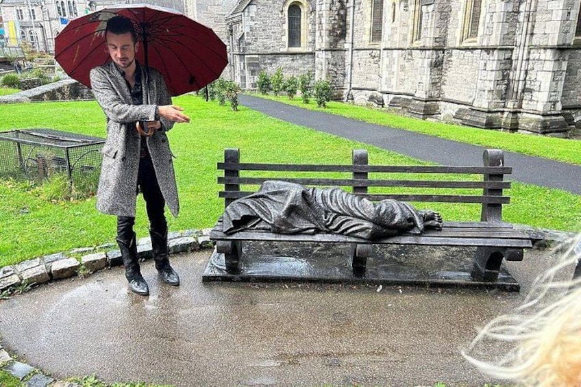 A person standing under an umbrella next to a bench with a statue of a sleeping figure. 2