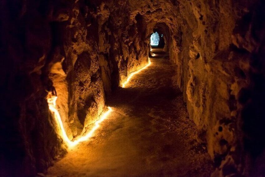 A narrow, illuminated cave passage with rough walls and a light at the end. 3