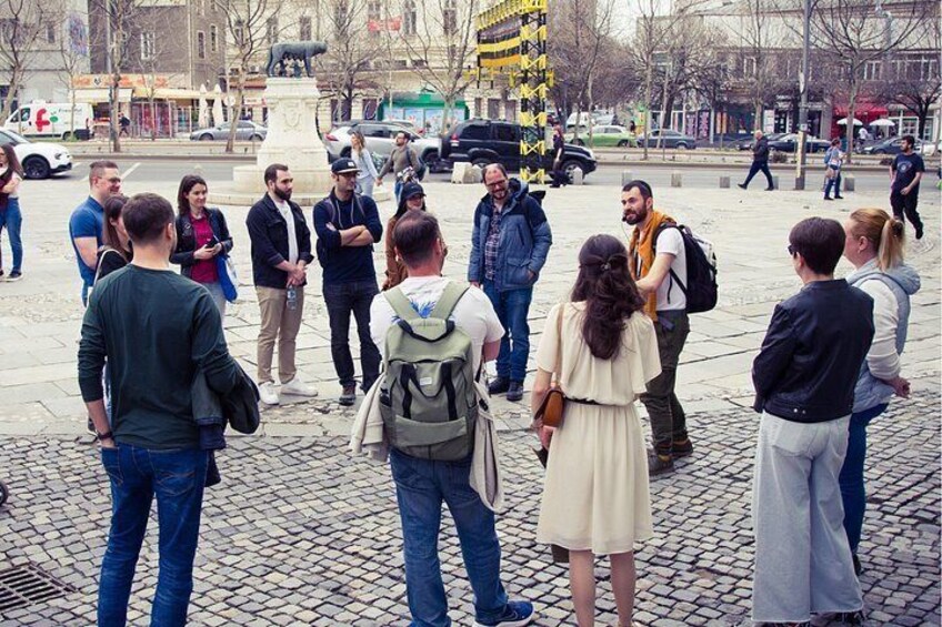 A group of people standing on a cobblestone street, a statue, and a busy urban environment with cars and buildings in the background. 4