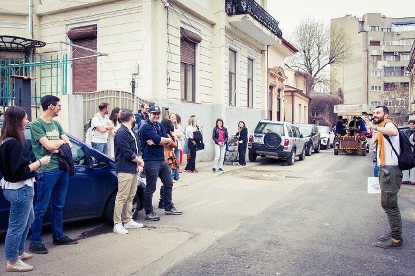 A street scene with people standing and walking, parked cars, and a building with a balcony. 5