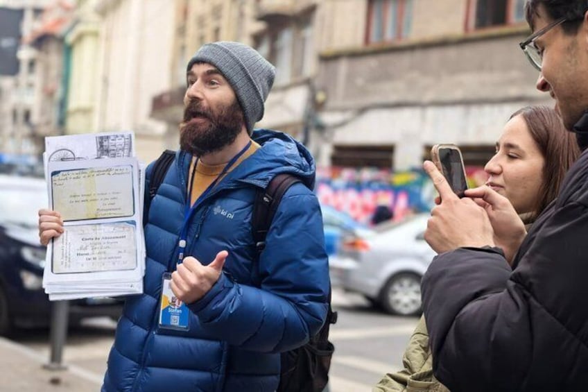 A person in a blue jacket holding documents, another person taking a photo, and a street with cars and buildings in the background. 1