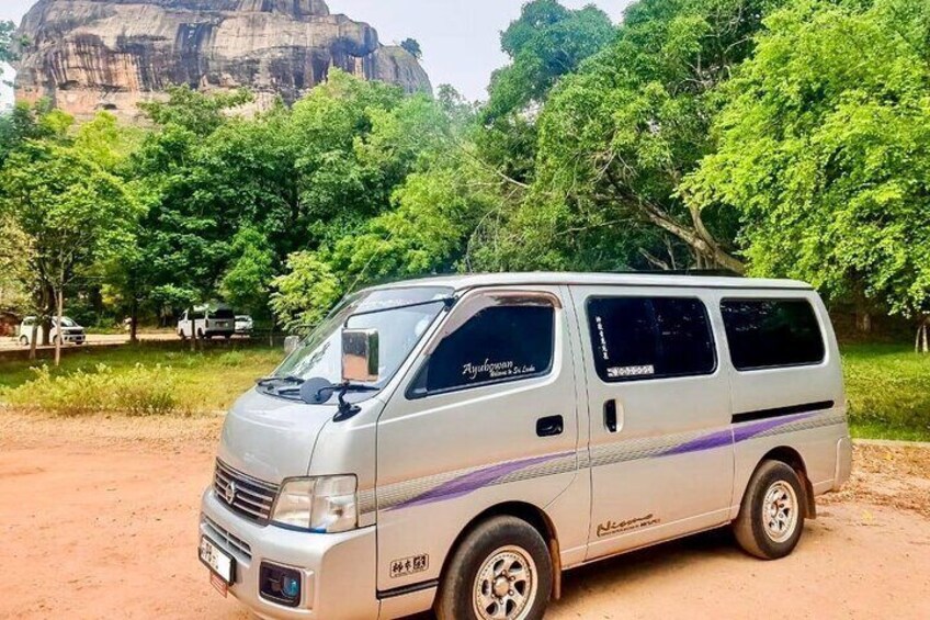 Una camioneta plateada estacionada en un camino de tierra, con árboles y una colina rocosa al fondo. 1