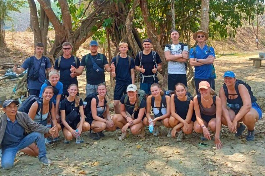 A group of people posing outdoors, some sitting and some standing, with trees and a dirt ground in the background. 4