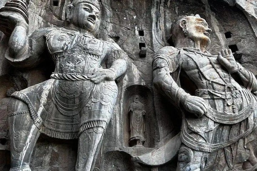 A stone relief of two armored figures, one seated and one standing, with intricate designs and a small inset figure. 3