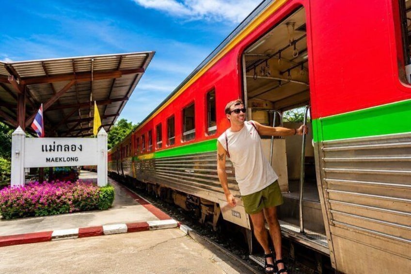 A train station platform with a red and green train, a person standing on the platform, and a sign with Thai script. 5