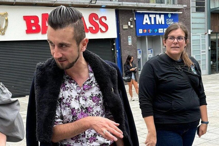 Un hombre con una camisa estampada y un abrigo negro caminando frente a una sucursal de Barclays y un cajero automático. 2